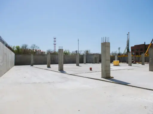 Plac budowy z betonową płytą i słupami betonowymi konstrukcyjnymi. W tle budynek i dźwig.