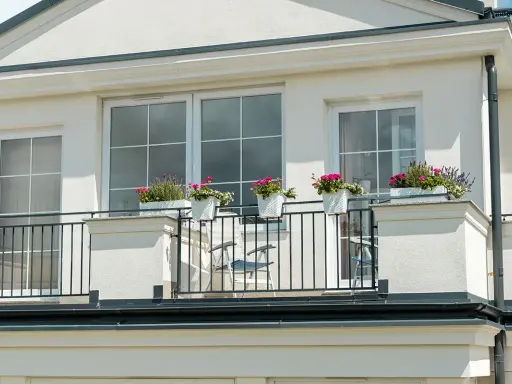 Białe okna pięknie komponują się z jasną, tynkowaną elewacją, tworząc elegancki wygląd. Balkon ozdobiony kwiatami dodaje uroku.