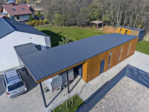 Nowoczesny budynek z drewnianą elewacją i dachem jednospadowym 7m. Obok stoi samochód i przyczepa pod zadaszeniem.