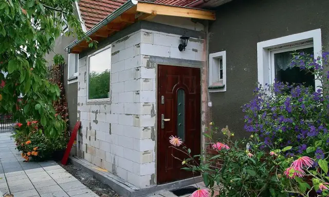 Rozbudowa domu na zgłoszenie: nowe wejście z białych pustaków, brązowe drzwi, okno i kwitnące kwiaty.