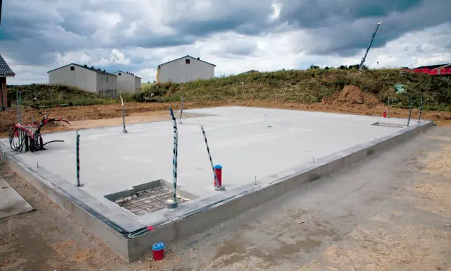 Budowa płyty fundamentowej w trakcie. Betonowa płyta z wystającymi rurami i kablami, w tle domy w budowie.