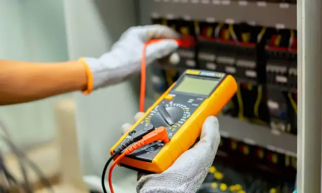 Elektryk w rękawicach wykonuje pomiar instalacji elektrycznej za pomocą żółtego multimetru.