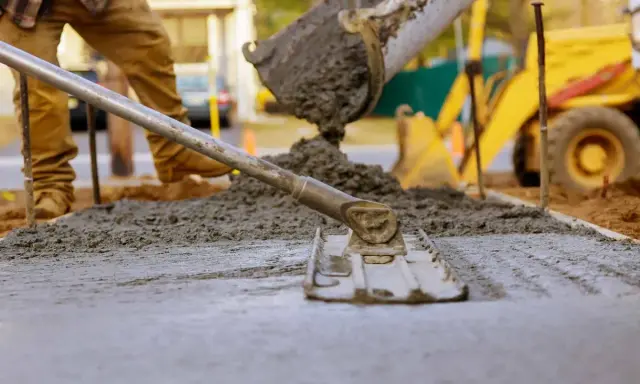 Pracownik wylewa beton, wyrównując go narzędziem. W tle koparka. Beton można lać do ok. 30°C.