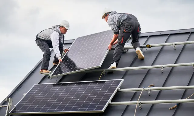 Dwóch monterów instaluje panele fotowoltaiczne na ciemnym, szczelnym dachu. Praca nad przyszłością energetyki.