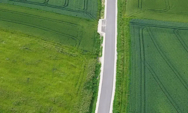 Wąska droga dojazdowa do działki, otoczona polami zielonych zbóż i traw.