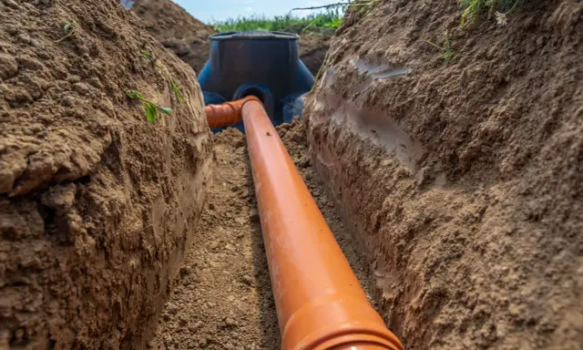 Wykonanie przyłącza wodociągowego: pomarańczowa rura biegnie przez wykop w ziemi do czarnej studzienki.
