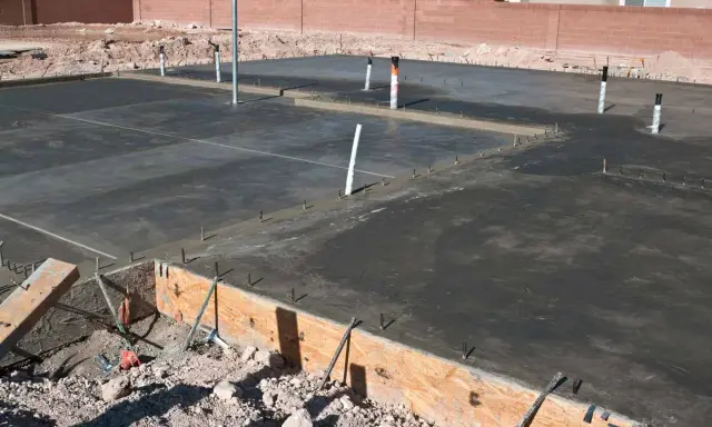 Świeżo wylany betonowy fundament z widocznymi rurami i zbrojeniem. Ziemia wokół czeka na materiał, czym obsypać fundamenty z zewnątrz.