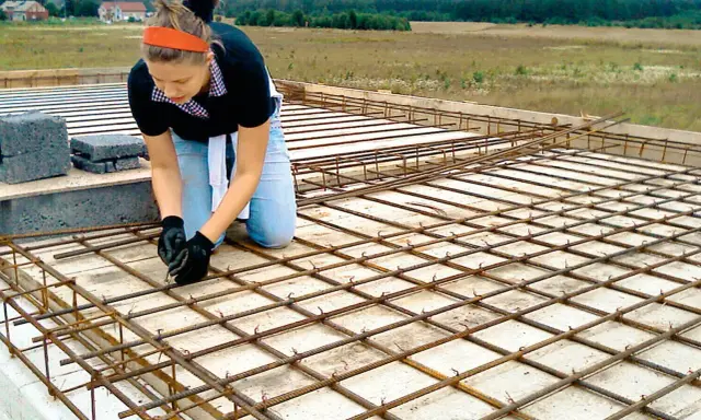 Kobieta w pomarańczowej opasce i jeansach klęczy, wiążąc zbrojenie. To element budowy domu samemu, gdzie każdy detal ma znaczenie.