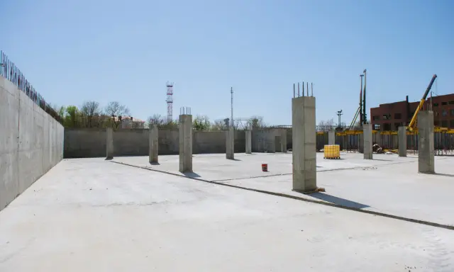 Plac budowy z betonową płytą i słupami betonowymi konstrukcyjnymi. W tle budynek i dźwig.