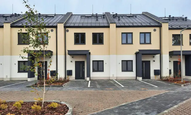 Nowe domy szkieletowe w zabudowie szeregowej, z jasnymi elewacjami i ciemnymi dachami. Przed domami parking i zieleń.