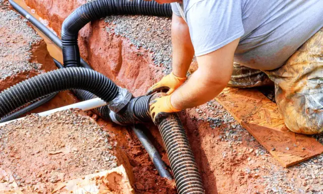 Instalator w pomarańczowych rękawiczkach łączy rury odprowadzające wodę z rynien. Prace zgodne z przepisami.