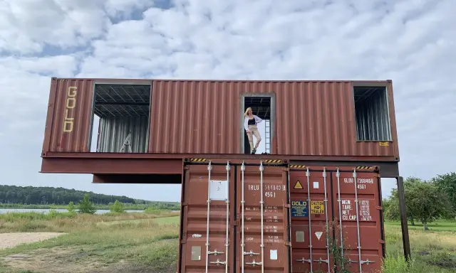 Dom z kontenerów w budowie. Kobieta stoi w otwartych drzwiach górnego kontenera, patrząc na krajobraz.