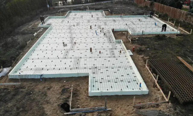 Szalunek płyty fundamentowej gotowy do zalania betonem. Na białej piance izolacyjnej ułożono zbrojenie i rury.