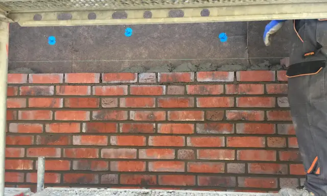 Budowa elewacji z klinkieru. Cegły układane na zaprawie, widoczna ręka w rękawicy i rusztowanie.