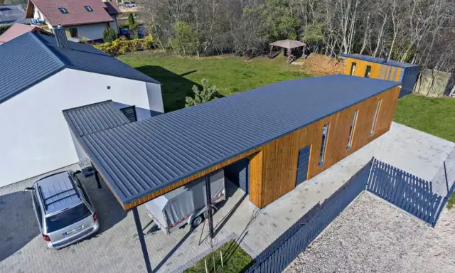 Nowoczesny budynek z drewnianą elewacją i dachem jednospadowym 7m. Obok stoi samochód i przyczepa pod zadaszeniem.