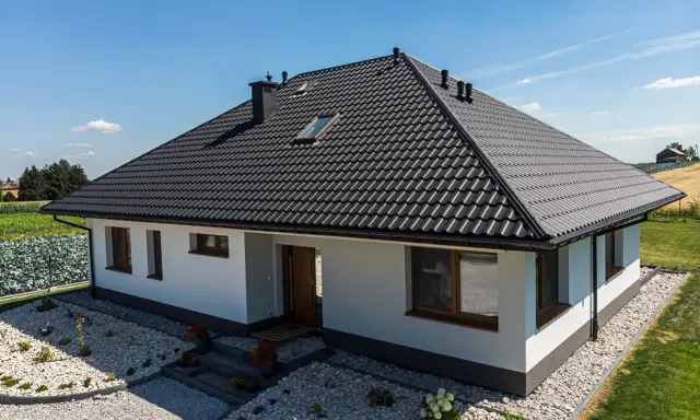 Nowy dom z czarnym dachem, gdzie kalenica jest najwyższym punktem dachu. Wokół zadbany ogród i pola.