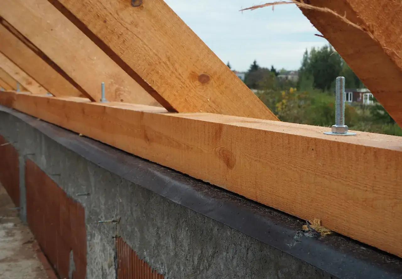 Murłata, czyli belka mocująca więźbę dachową, jest przykręcana do ściany. Widać drewniane krokwie i betonowy fundament.