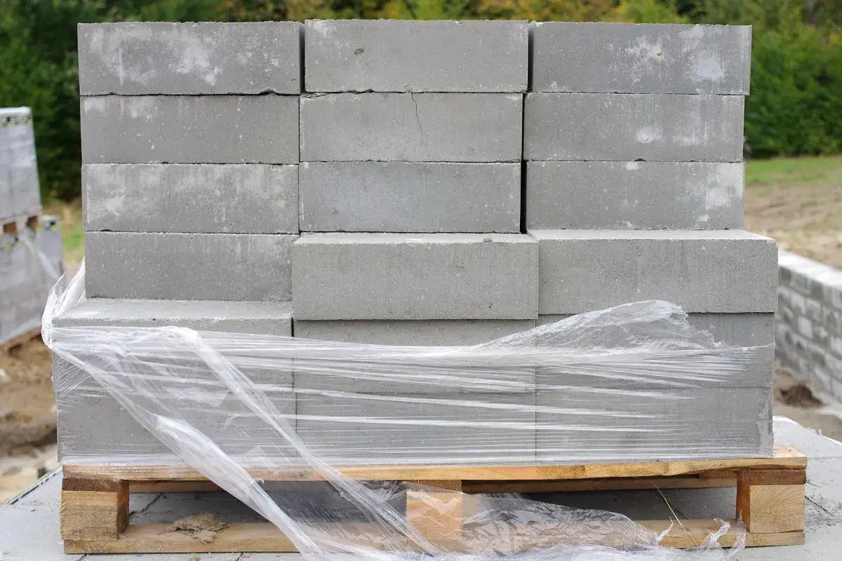 Stos szarych bloczków betonowych na palecie, owinięty folią. Poznaj bloczek betonowy wymiary dla swojej budowy.