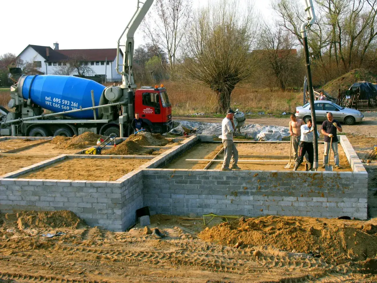 Budowa fundamentów: koparka, betoniarka i ekipa budowlana przygotowują teren. Czym obsypać fundamenty z zewnątrz? Piaskiem i ziemią.