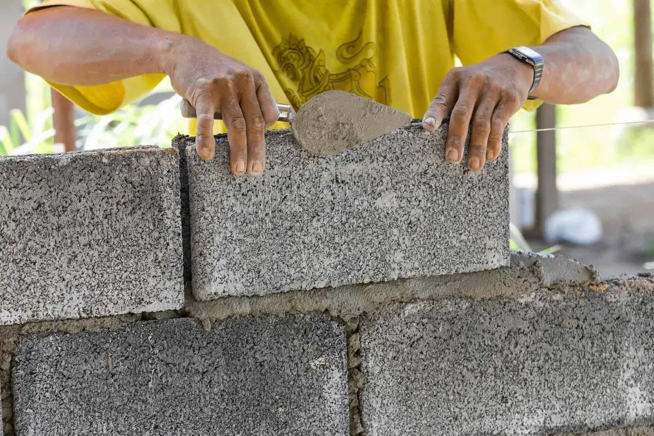 Ręce budowlańca układają betonowy bloczek na zaprawie. Trwa murowanie z bloczków betonowych.