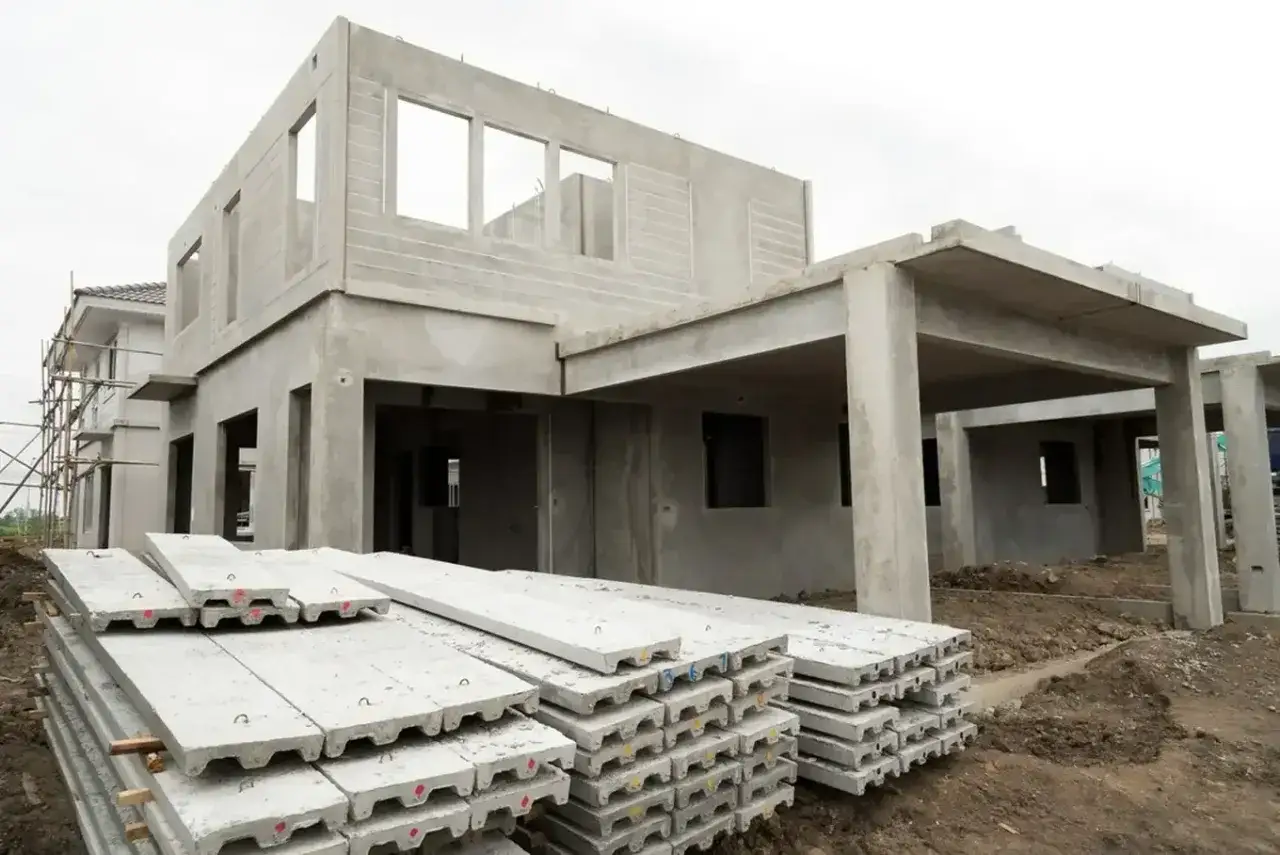 Budowa domu z prefabrykatów betonowych w toku. Na pierwszym planie leżą ułożone płyty betonowe, a w tle widać konstrukcję budynku z otworami okiennymi i drzwiami.
